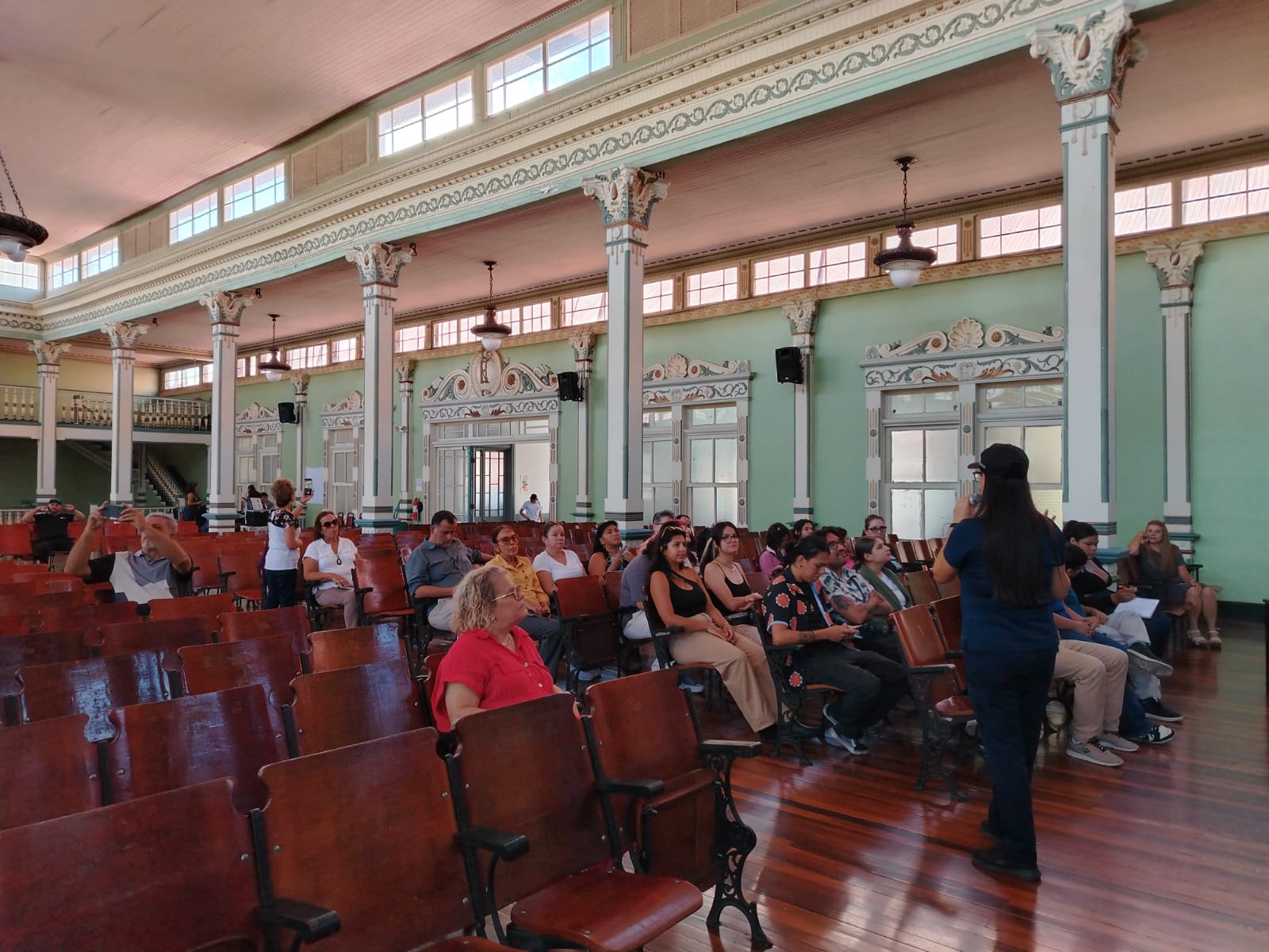 Personas sentadas en un salón antiguo con sillas de madera, una mujer al frente explicando en un micrófono.