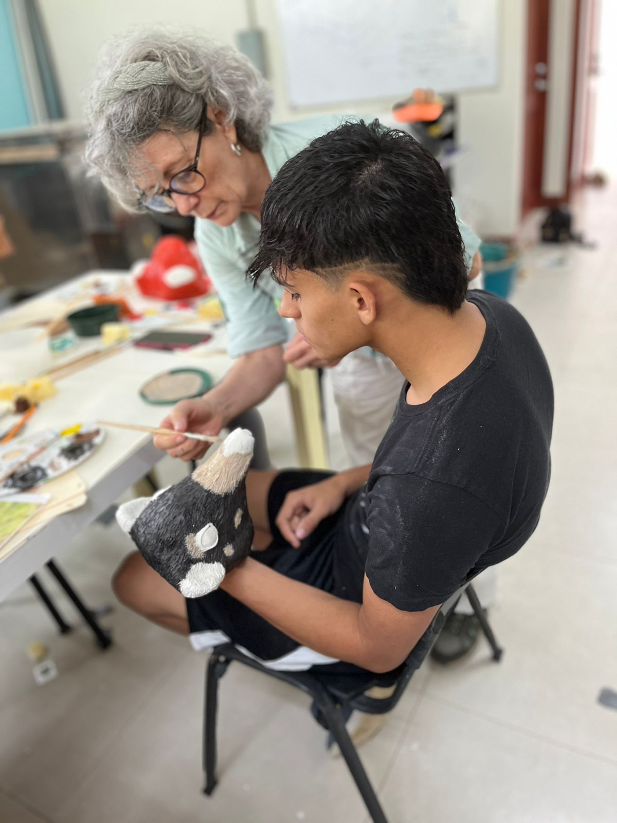 Una señora de pie pintando un objeto que tiene un joven en su mano.
