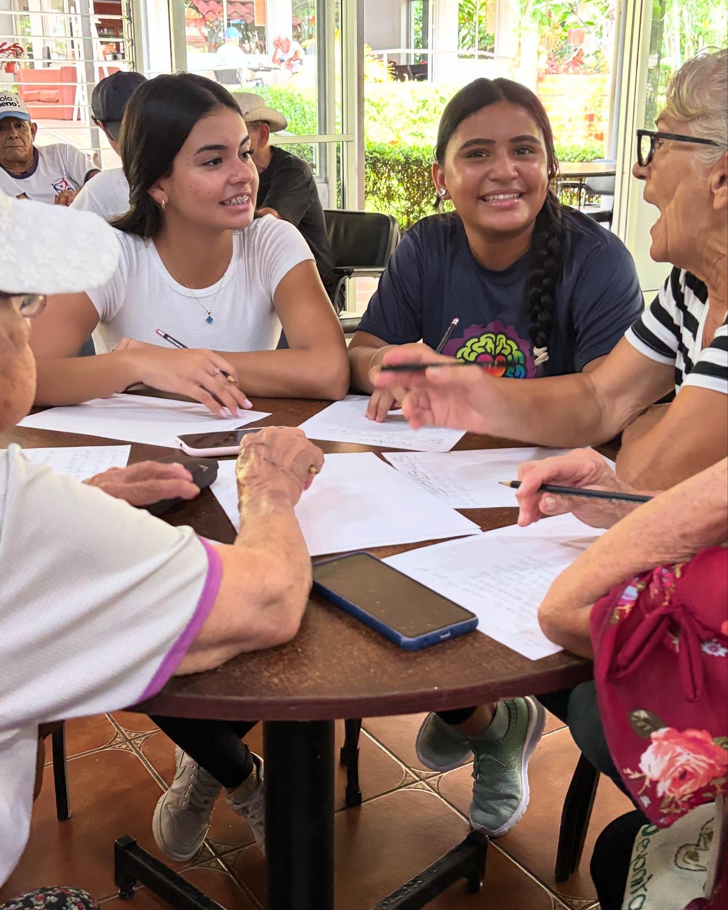 Dos mujeres jóvenes sonriendo y conversando con dos señoras mayores, todas sentadas en una mesa redondo con papeles en la mesa