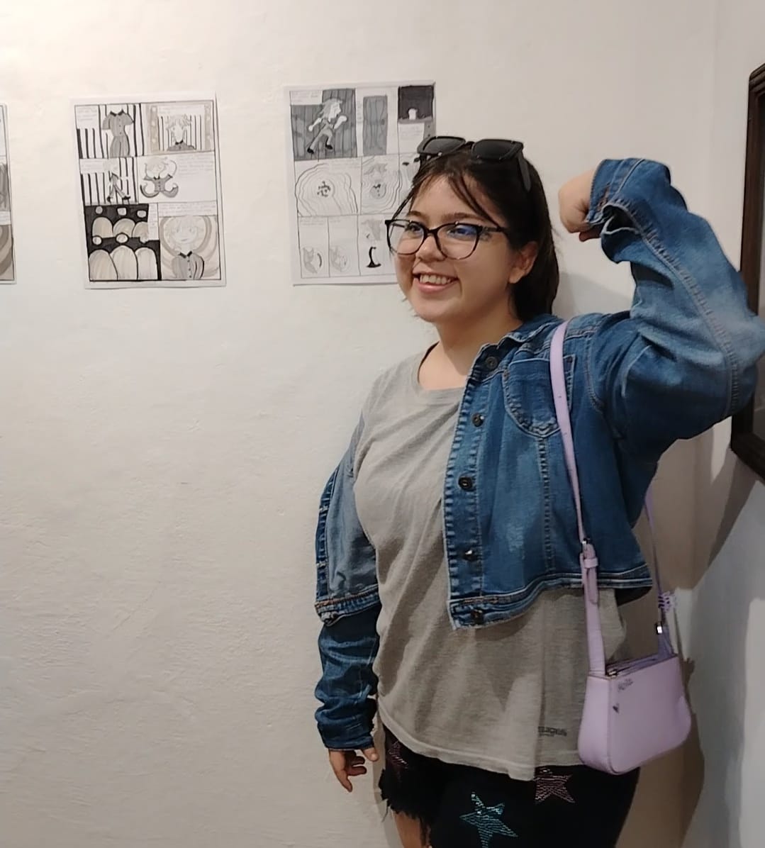 Una joven haciendo una pose de fuerza con el brazo, atrás se encuentran unos comics pegados a la pared.