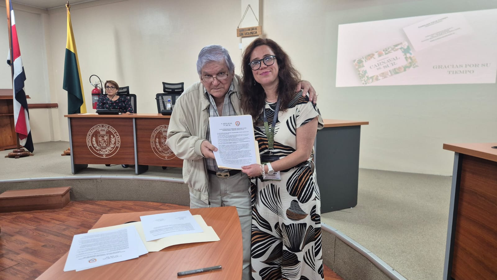Un hombre y una mujer sosteniendo un documento mirando hacia la cámara, al fondo una señora sentada en una mesa