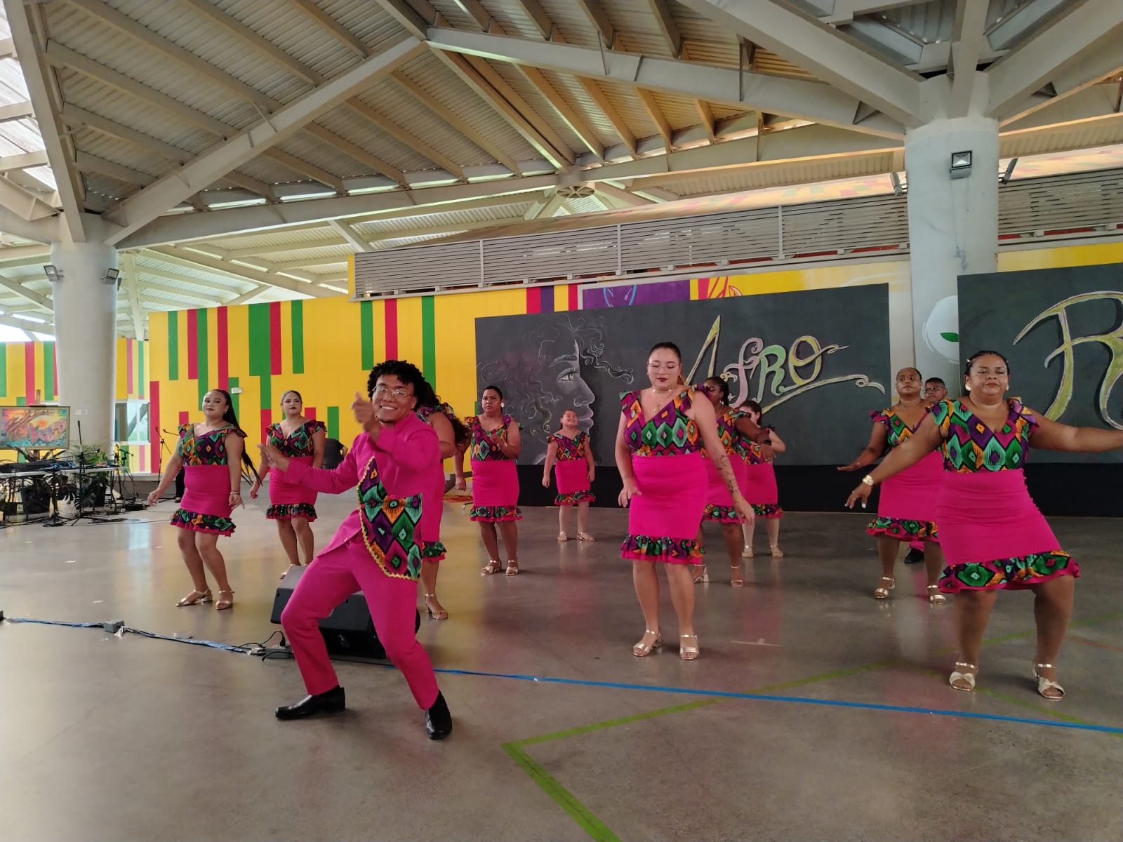 Un hombre al frente y varias mujeres distribuidas hacia atrpas vestidas con ropa afrocaribeña color rosa, bailando
