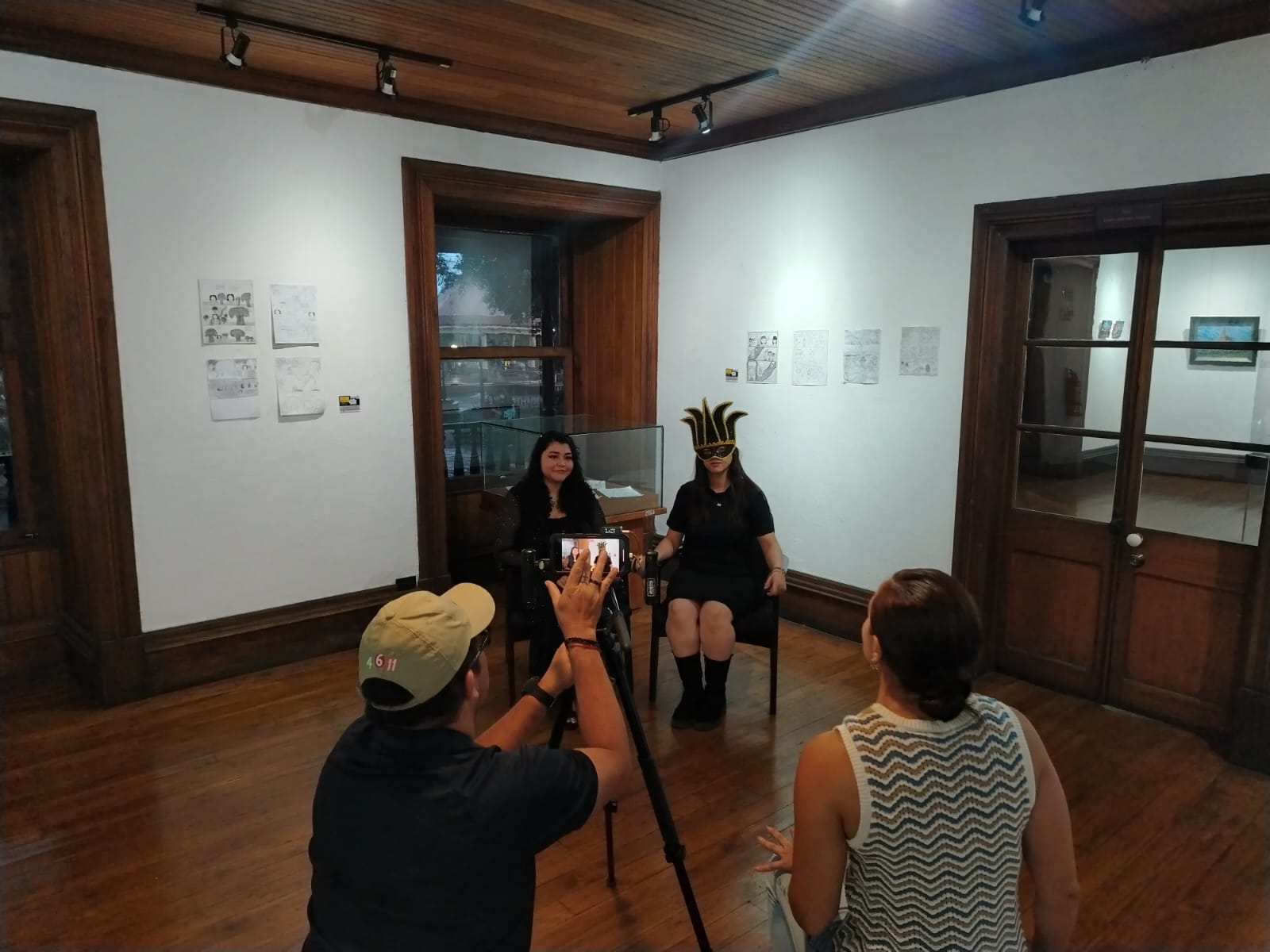 Dos mujeres jóvenes sentadas, una de ellas con antifaz. Al frente una mujer entrevistándolas y un hombre ajustando una cámara.