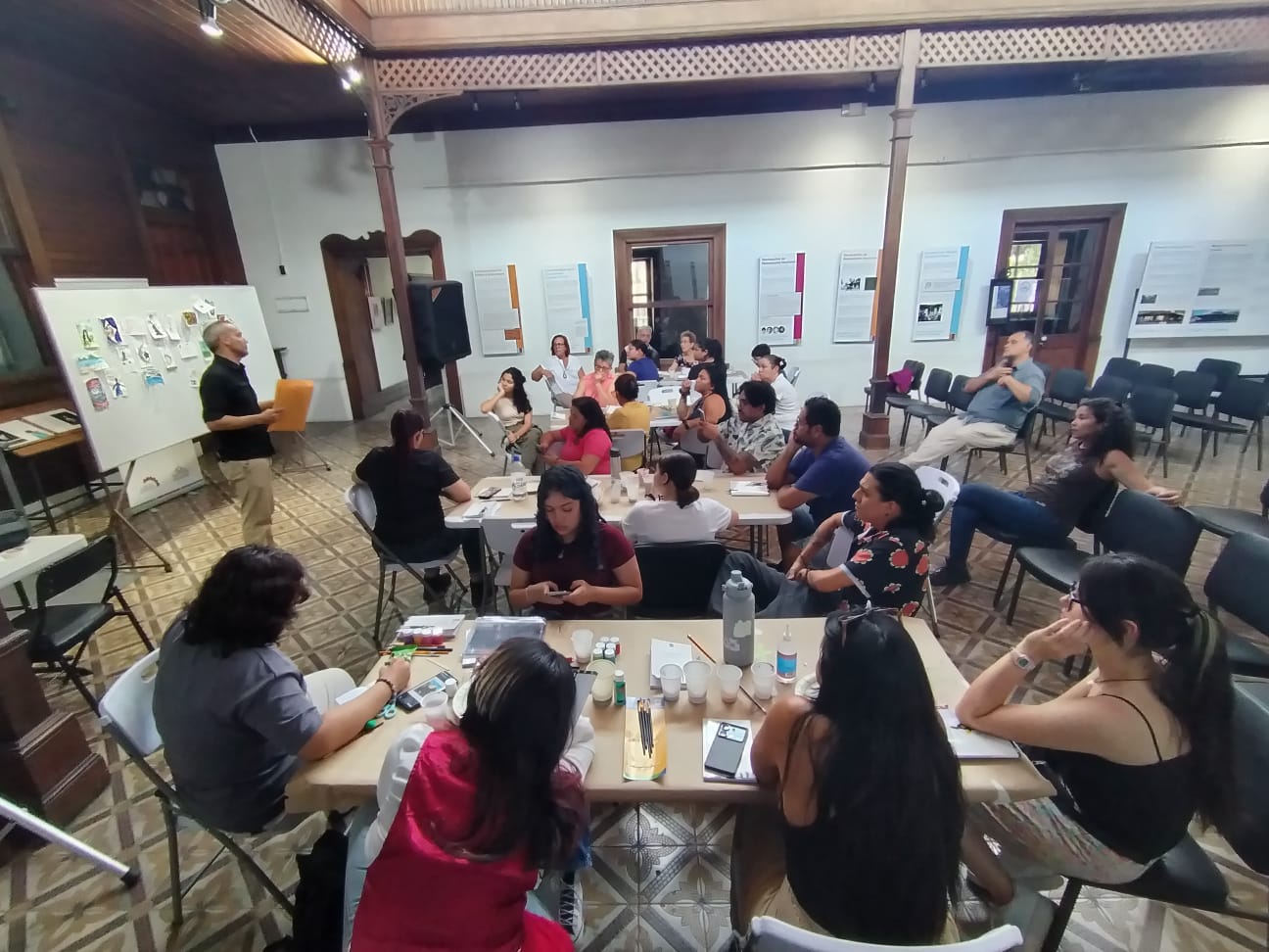 Grupos divididos en mesas con materiales de pintura, al frente un hombre explicando y otras personas sentadas.
