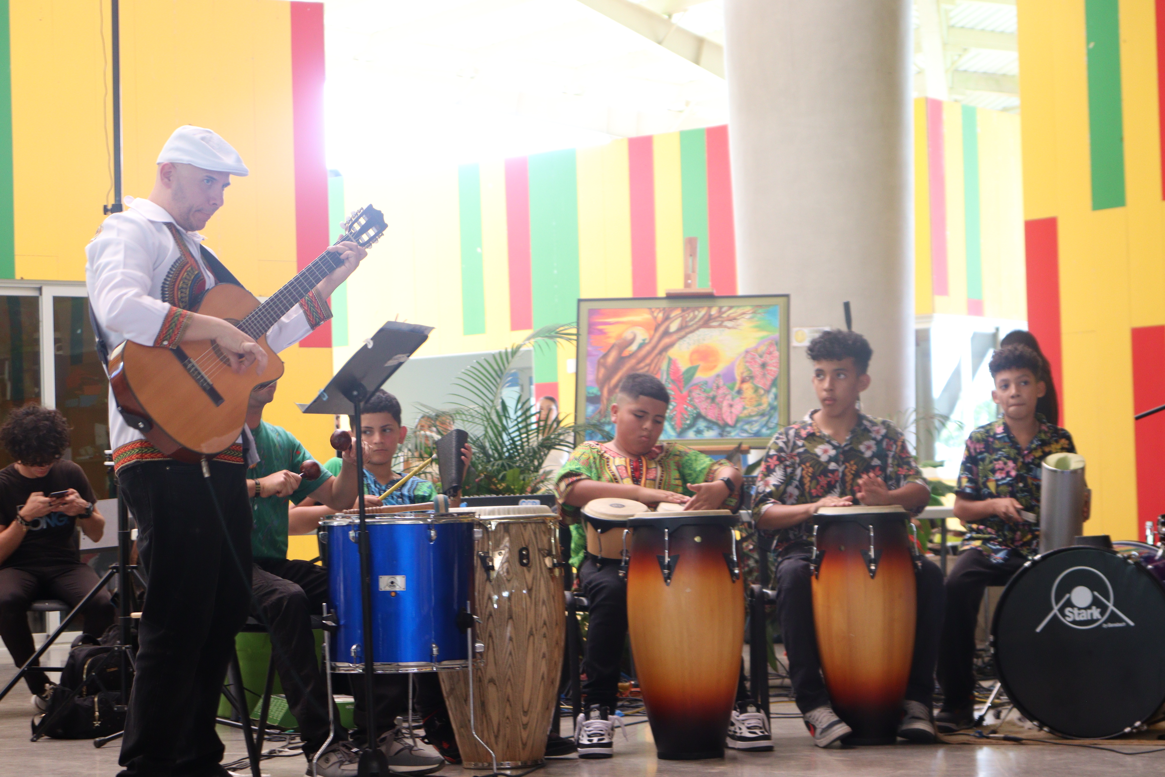 Hombre tocando una guitarra, niños tocando otros instrumentos como el güiro, timbales y batería.
