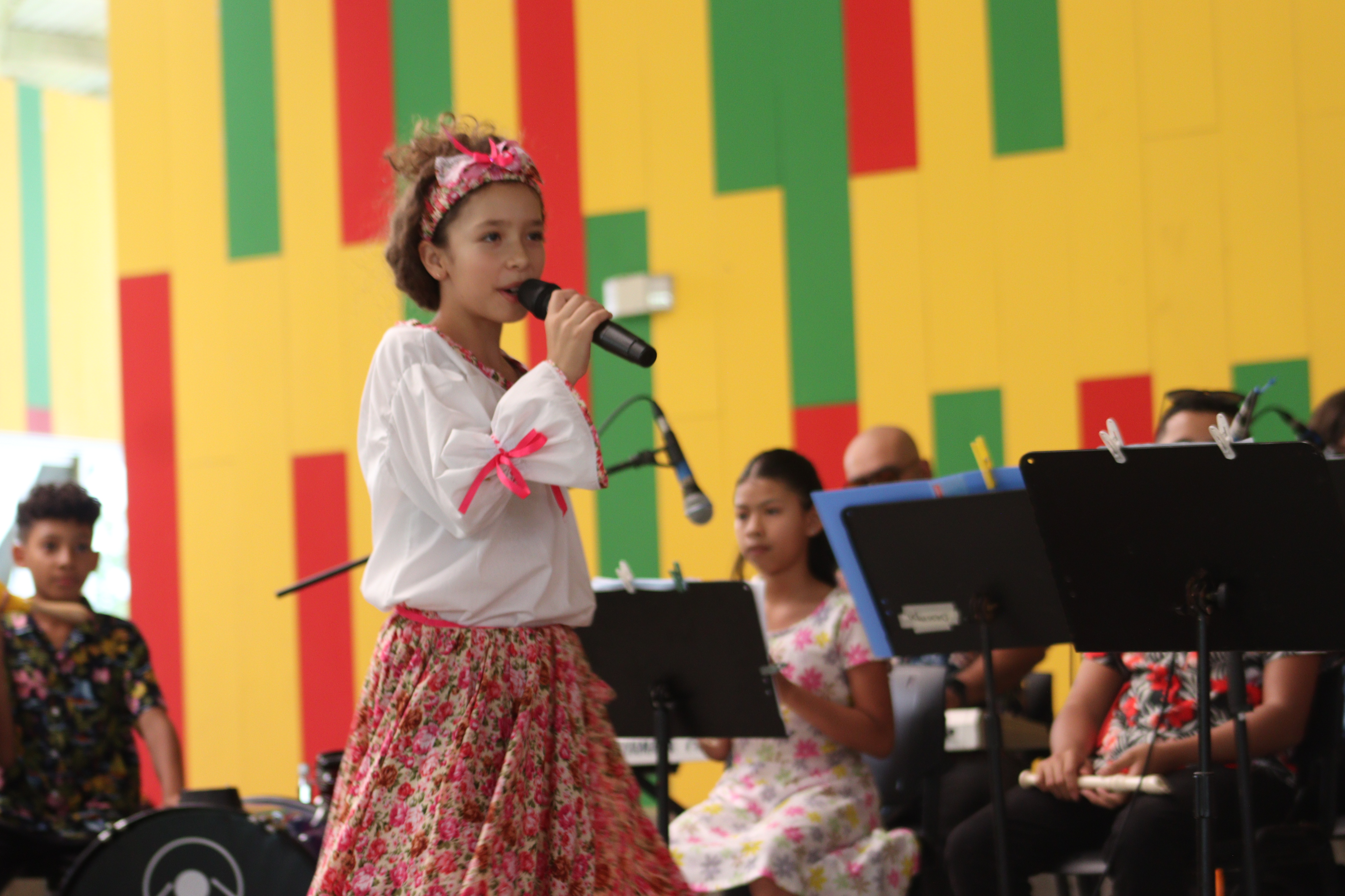 Mujer joven viste enagua floreada, blusa blanca y pañuelo en la cabeza, canta con un microfono. Atrás niños tocan instrumentos.