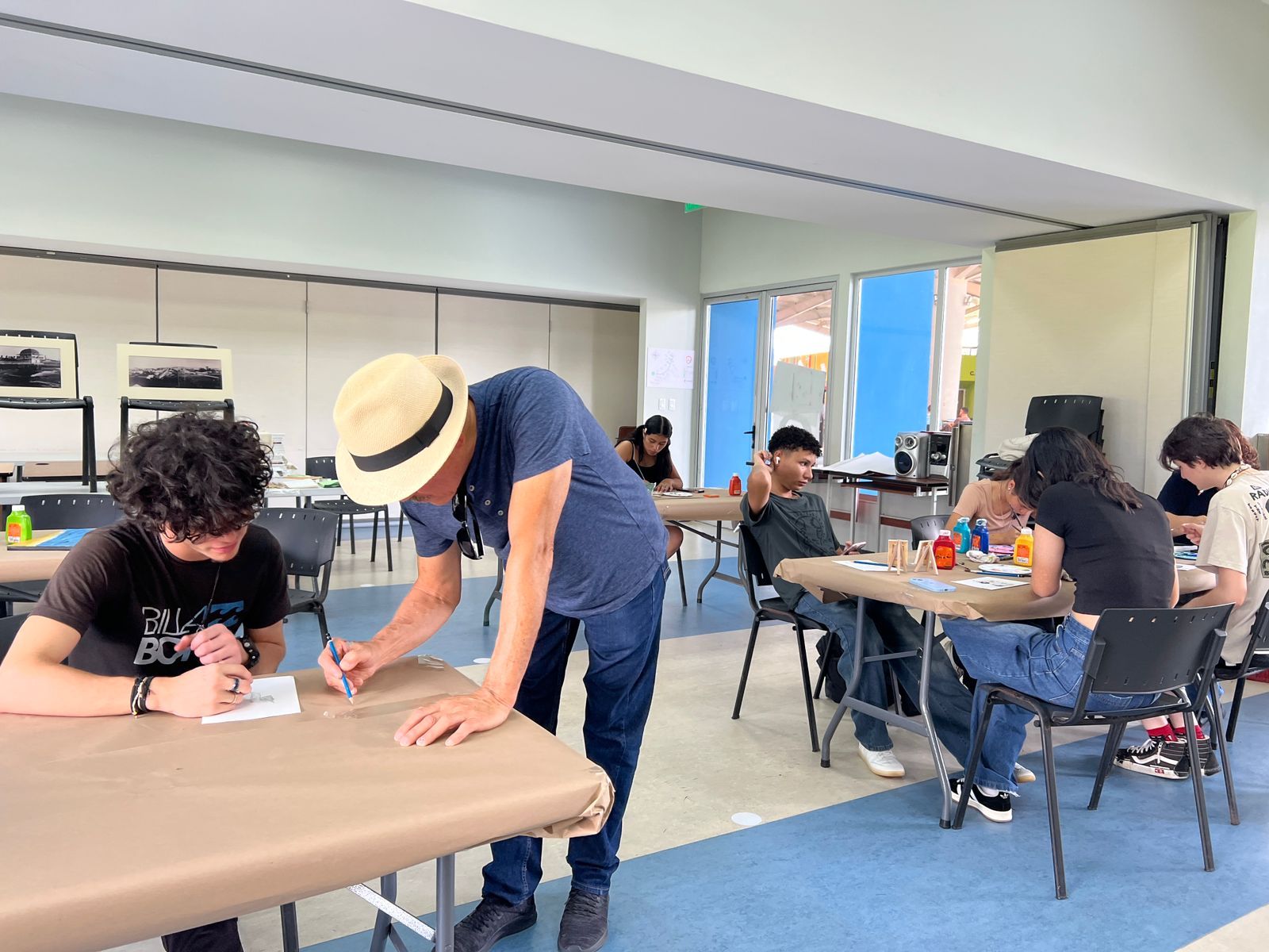 En un aula, un hombre con sombrero dibuja en una papel sobre la mesa frente a un joven que observa. Atrás un grupo de jóvenes sentados en una mesa