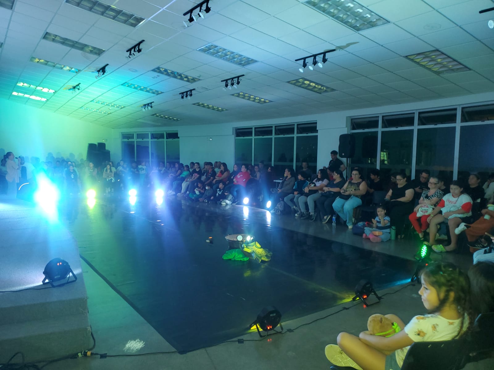 Personas sentadas alrededor de un escenario con piso negro y luces.