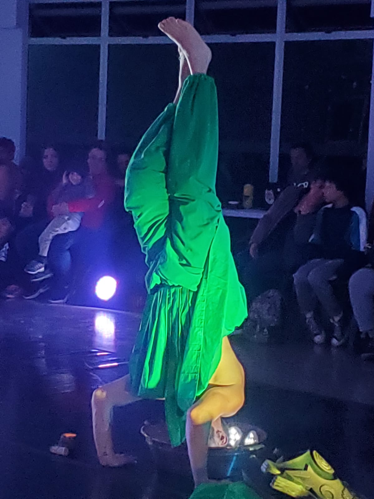 Un hombre de cabeza, vestido de verde, metiendo su cabeza en un balde. Publico al fondo.