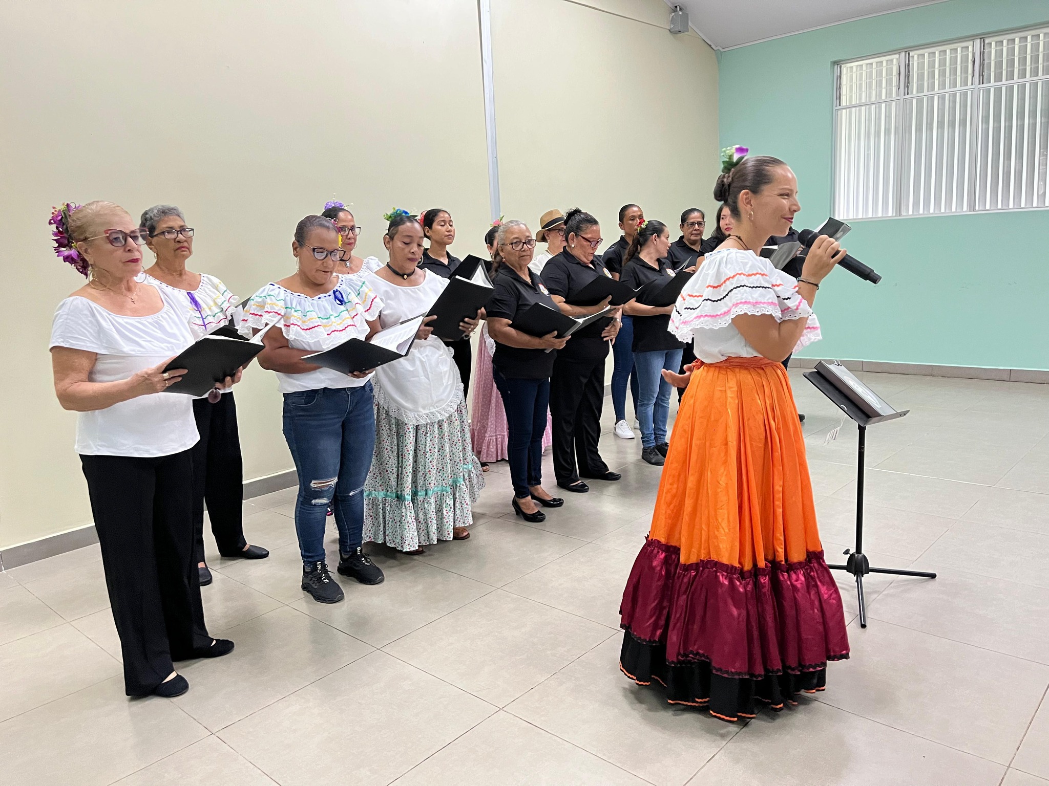 Mujer con vestido tipico sujetando un micrófono, y atrás señoras de pie sosteniendo un cuaderno negro.