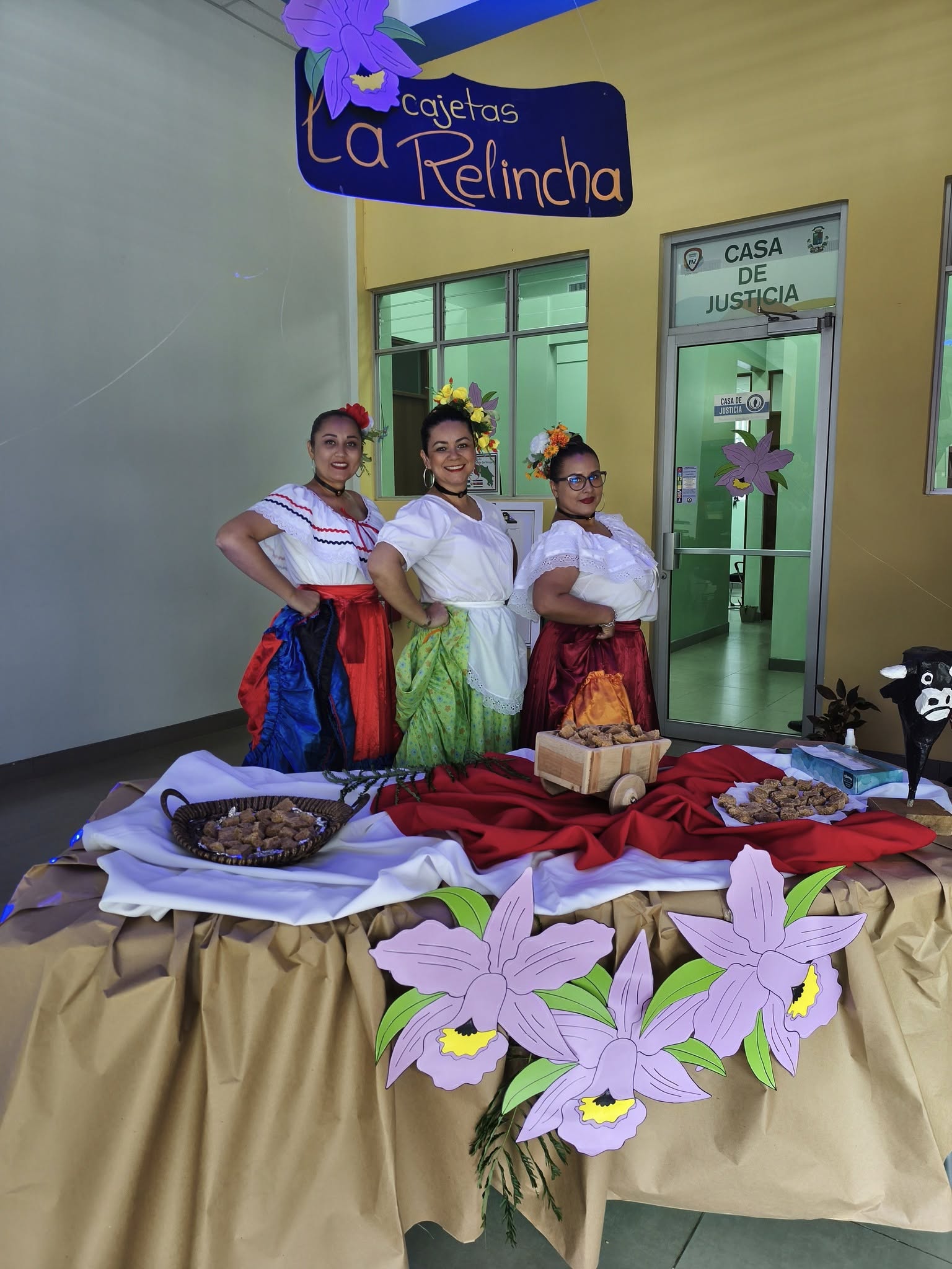 Una mesa con unas guarias moradas de papel al frente y reposterías encima, atrás tres mujeres posando con traje tipico y arriba un letrero que dice Cajetas La Relincha