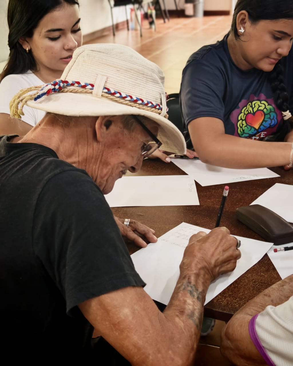Un señor con sombrero blanco escribiendo en una hoja con un lápiz. Atrás dos mujeres jóvenes escribiendo en un papel