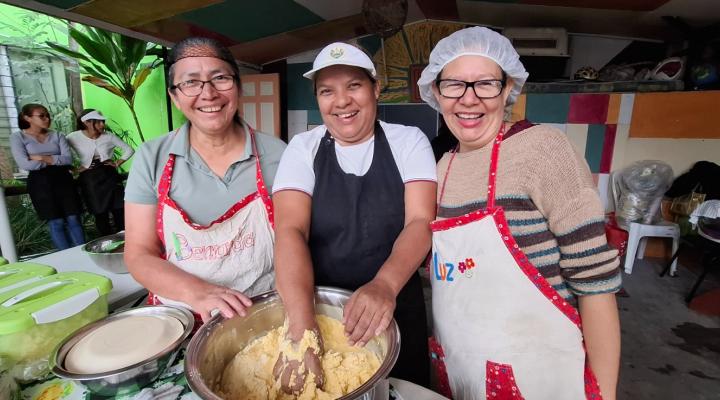 Tres mujeres con delantal y gorro de cocina, sonriendo. Una de ella con su mano en una mezcla de masa.