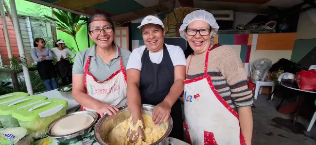 Tres mujeres con delantal y gorro de cocina, sonriendo. Una de ella con su mano en una mezcla de masa.