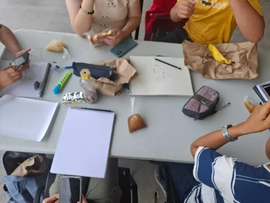 Una mesa con lapices, hojas en blancos y cuadernos y personas alrededor